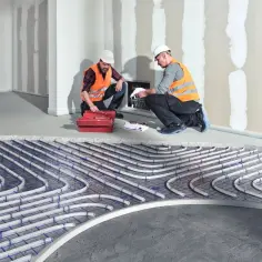 Dwóch budowlańców w kamizelkach odblaskowych sprawdza projekt instalacji podłogowego ogrzewania. Widać rurki ułożone na styropianie, który jest kluczowy jako izolacja.