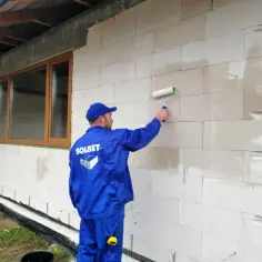 Pracownik w niebieskim uniformie nakłada wałkiem grunt do styropianu na ścianę z bloczków.