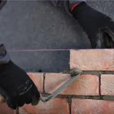 Ręce w czarnych rękawicach wykonują murowanie ścian działowych, układając cegły na zaprawie.