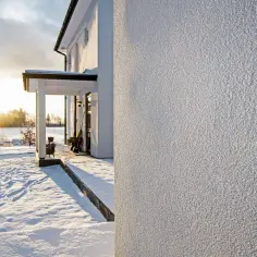 Śnieg na podwórku i zachodzące słońce podkreślają fakturę świeżo nałożonego tynku silikonowego na ścianie domu.