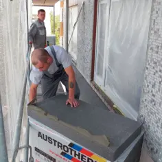 Budowlaniec układa styropian pod wylewkę na balkonie. Obok stoi drugi pracownik.