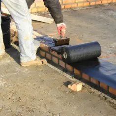 Pracownik nakłada masę hydroizolacyjną do fundamentów na ceglaną ścianę, przygotowując podłoże pod izolację.