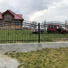 Nowoczesne płoty kute z ozdobnymi elementami, otaczające posesję z domem i czerwonym samochodem.