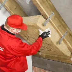 Pracownik w czerwonym kombinezonie montuje wełnę mineralną, wykonując ocieplenie murłaty od wewnątrz.