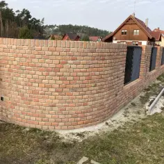 Eleganckie ogrodzenie z klinkieru, z łukiem i metalowymi wstawkami, otacza posesję z domami w tle.