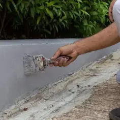 Malowanie betonowego murku farbą do betonu na zewnątrz. Ręka z pędzlem pokrywa szarą farbą szary murek.