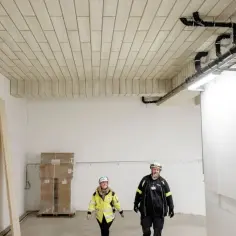Ocieplenie sufitu w garażu z widocznymi rurami i instalacjami. Dwie osoby w kaskach inspekcjonują postęp prac.