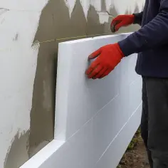 Pracownik w czerwonych rękawicach montuje płyty styropianowe na ścianie. To etap prac związanych z tym, ile kosztuje ocieplenie budynku.