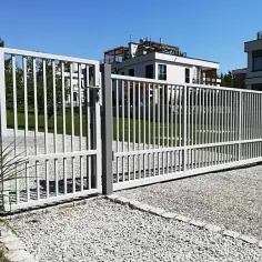 Brama przesuwna na pilota, biała, z pionowymi szczebelkami, otwiera wjazd na posesję z nowoczesnymi budynkami.