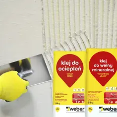 Ręka w żółtej rękawicy nakłada klej na styropian. Obok worki z klejem do ociepleń i wełny mineralnej Weber.