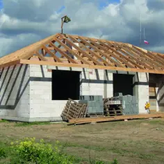 Budowa domu z widocznym szkieletem dachu dwuspadowego. Materiały budowlane leżą na placu.