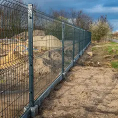 Trwa montaż ogrodzenia panelowego. Nowy płot z betonową podmurówką oddziela plac budowy od drogi i terenów zielonych.