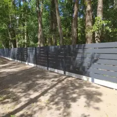 Nowoczesne, ciemnoszare ogrodzenie z poziomymi szczelinami, osadzone na betonowym cokole, na tle lasu.