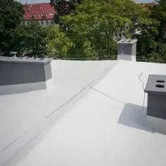 Nowy dach z białą hydroizolacją na papę, z szarymi kominami i zielenią w tle.