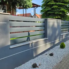 Nowoczesny płot beton architektoniczny z szarymi, poziomymi przęsłami. Ozdobne kamyczki i miniaturowe krzewy tworzą schludny ogród.