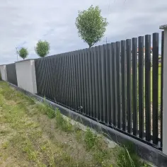 Nowoczesne ogrodzenia panelowe na nierównym terenie, z betonowymi słupkami i ciemnoszarymi panelami, tworzą solidną barierę.