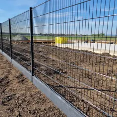 Solidne, tymczasowe ogrodzenie placu budowy zabezpiecza teren. W tle widać materiały budowlane i domy.