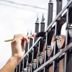 Ręka maluje pędzlem ozdobną, metalową bramę. Używana jest farba chlorokauczukowa do metalu, która chroni przed rdzą.
