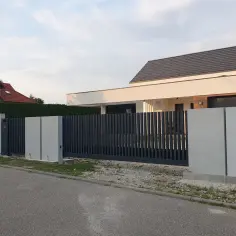 Nowoczesne ogrodzenie z beton architektoniczny i ciemnoszarymi panelami, otaczające posesję z domem.