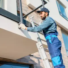 Pracownik na drabinie montuje balustradę na balkonie. Widać styrodur na balkonie bez wylewki, przygotowany do dalszych prac.