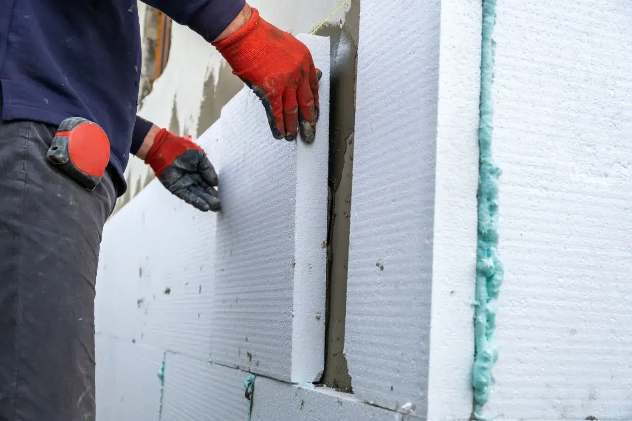 Ręce w czerwonych rękawiczkach przykładają płyty styropianu do ściany. Widać zaprawę i piankę montażową, co pokazuje, jak kleić styropian.