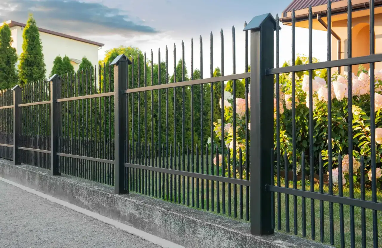 Betonowy murek jako alternatywa dla podmurówki pod ogrodzenie. Czarny płot z ostrymi zakończeniami, za nim tuje i kwitnące krzewy.