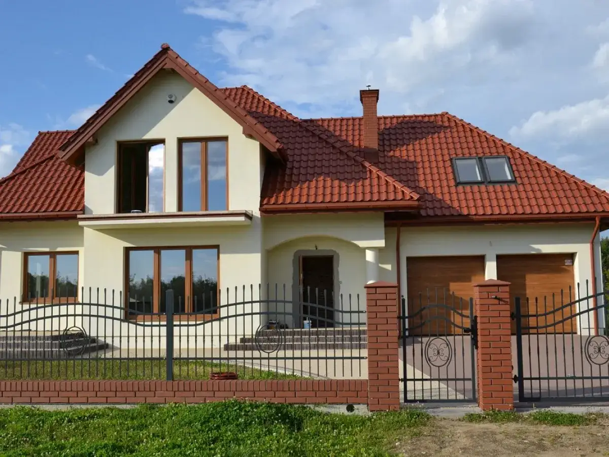 Eleganckie ogrodzenia z klinkieru wzory, z metalowymi przęsłami i ozdobnymi słupkami, otaczają nowoczesny dom z czerwoną dachówką.