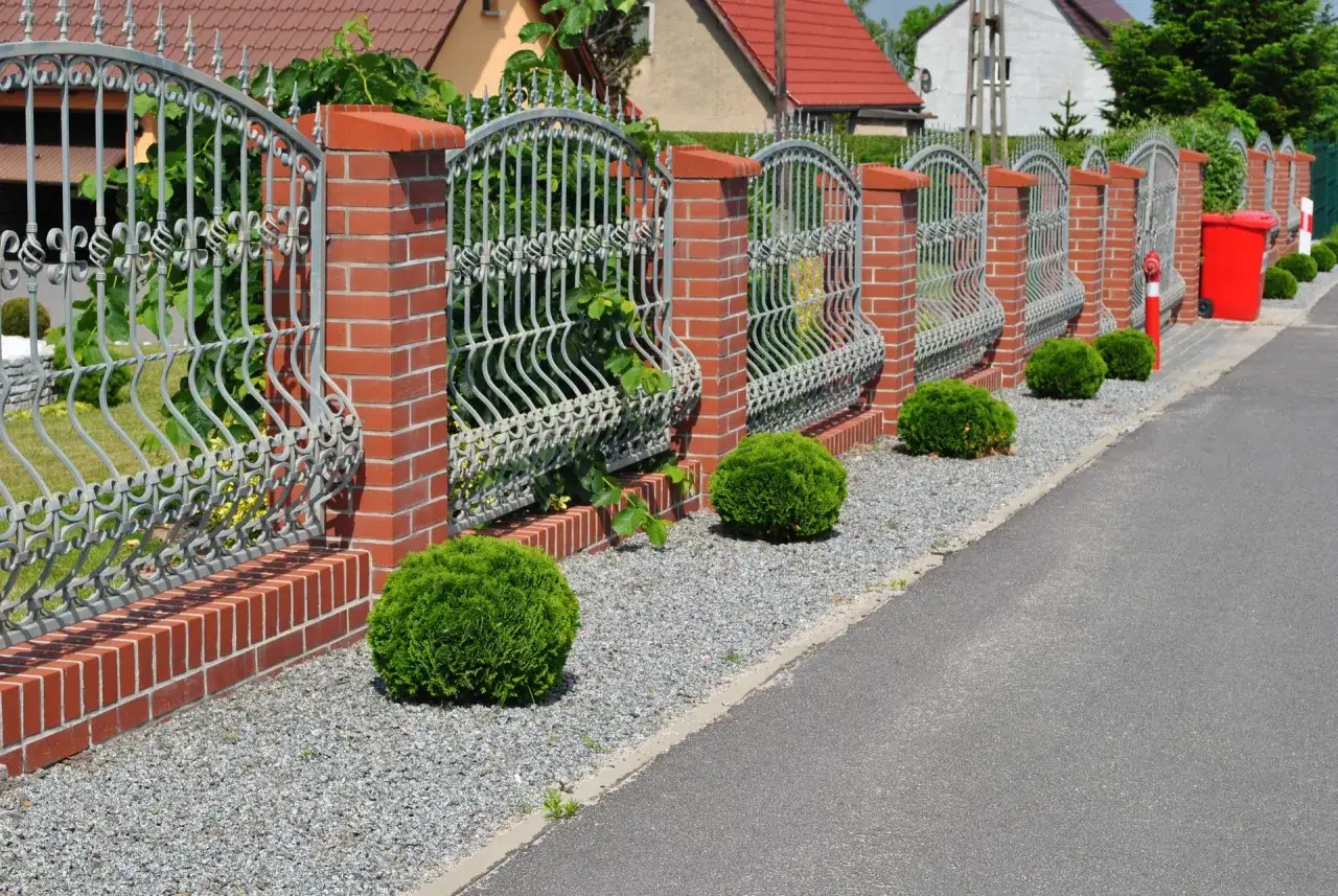 Piękny, kuty płot z ceglanymi słupkami wyznacza, która strona płotu należy do właściciela. Krępa zieleń i żwir tworzą schludny ogród.