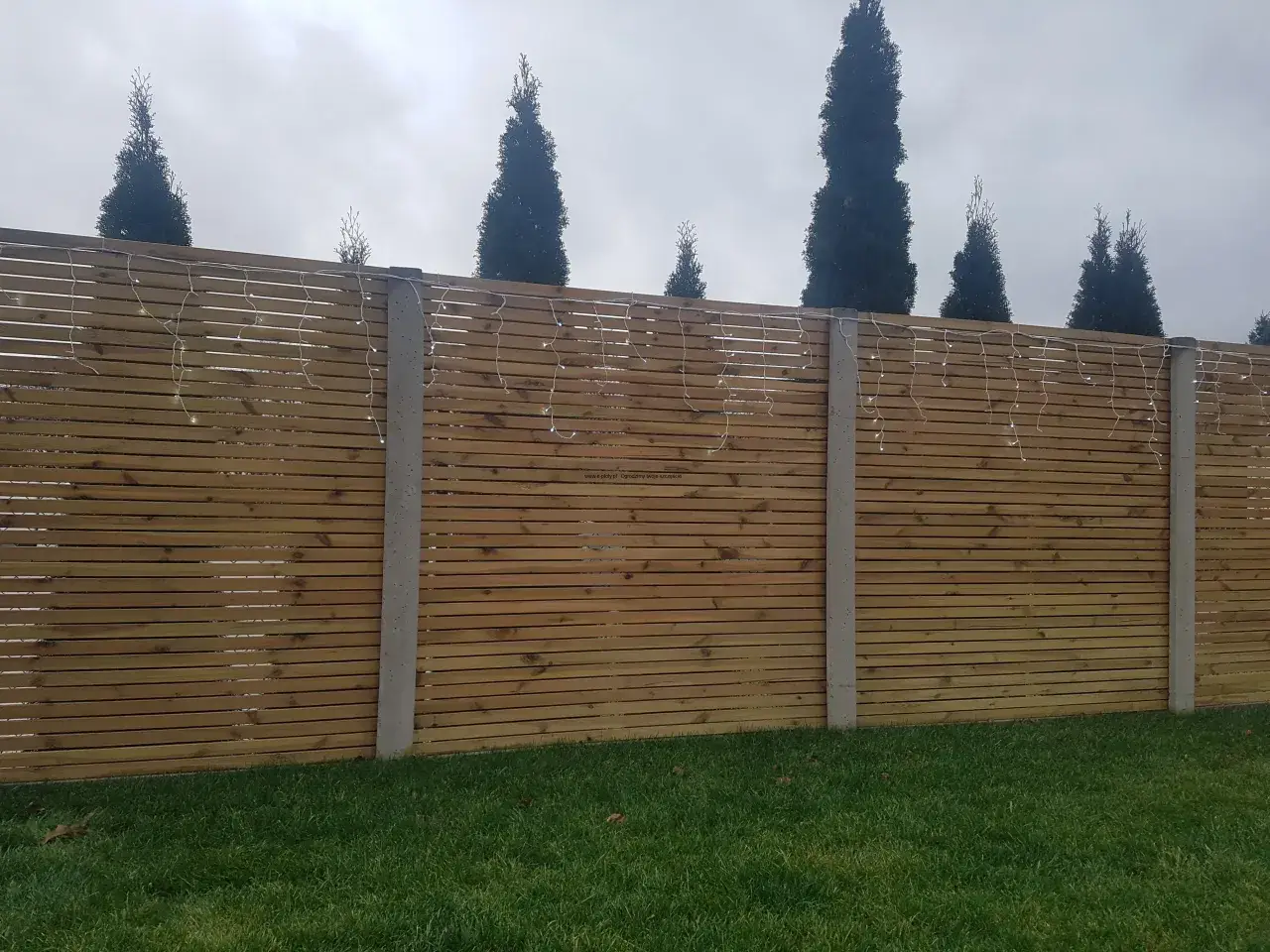 Drewniane deski i betonowe słupy tworzą nowoczesne ogrodzenie. Girlanda świetlna dodaje uroku, stanowiąc pomysł na tanie ogrodzenie działki.