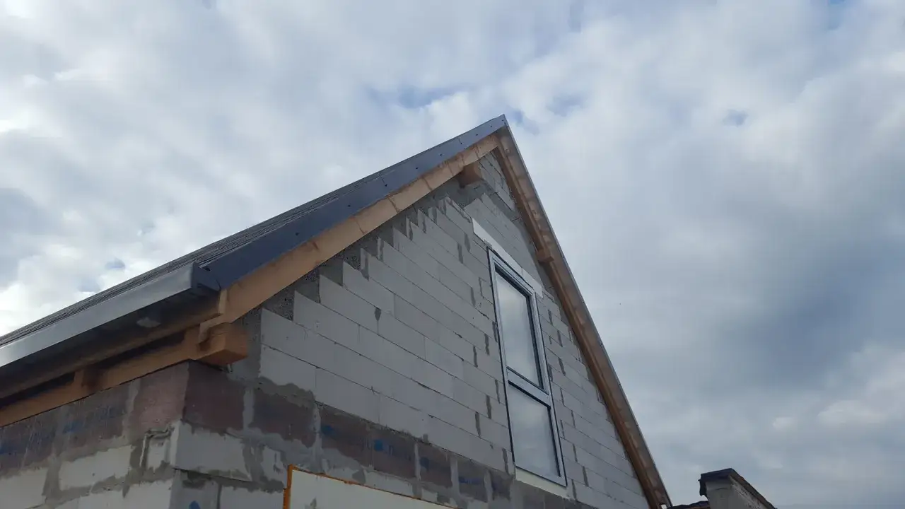 Rozpoczyna się wykończenie ścian szczytowych domu. Widać szary beton komórkowy, drewniane elementy dachu i okno.