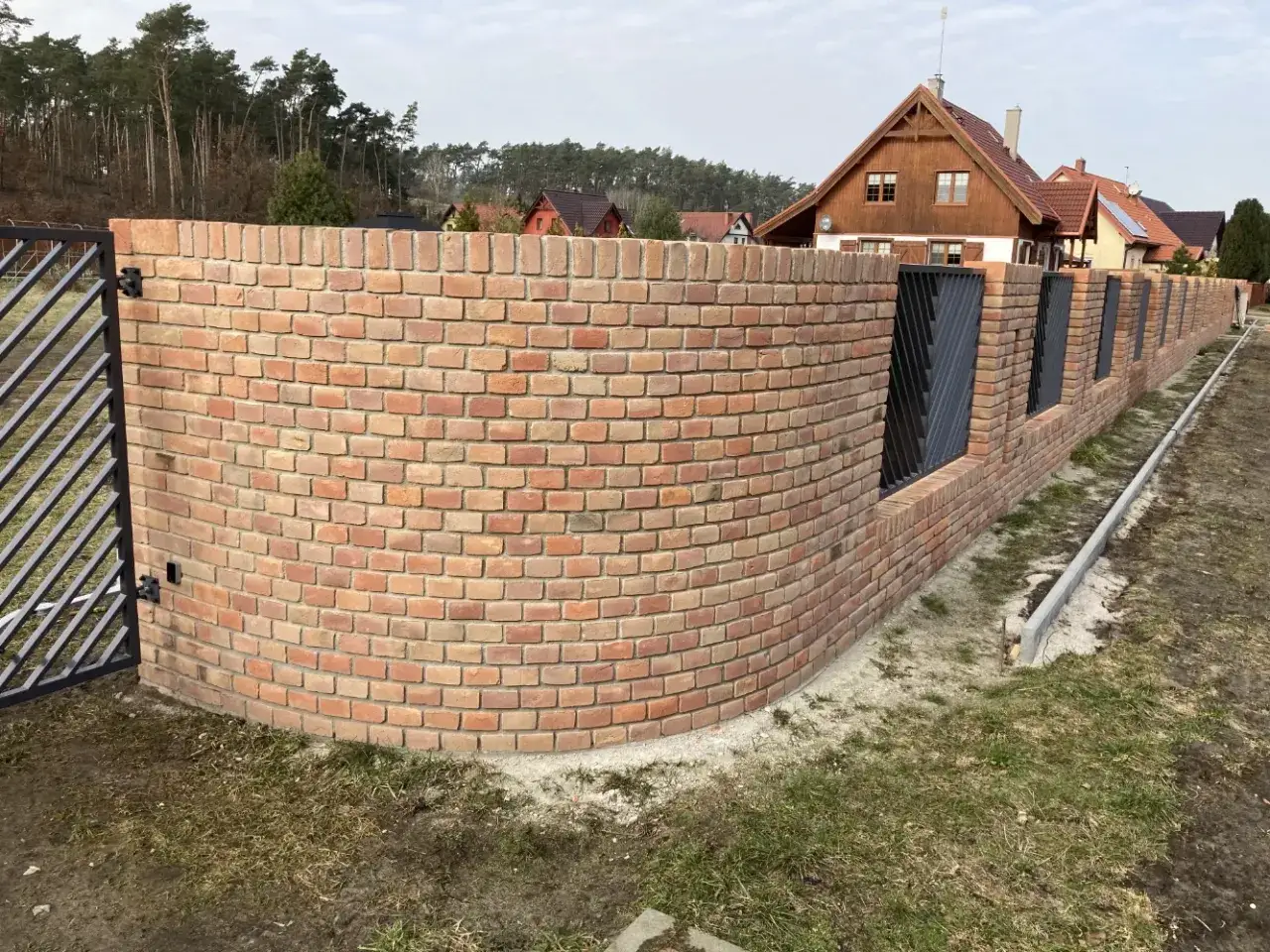 Eleganckie ogrodzenie z klinkieru, z łukiem i metalowymi wstawkami, otacza posesję z domami w tle.