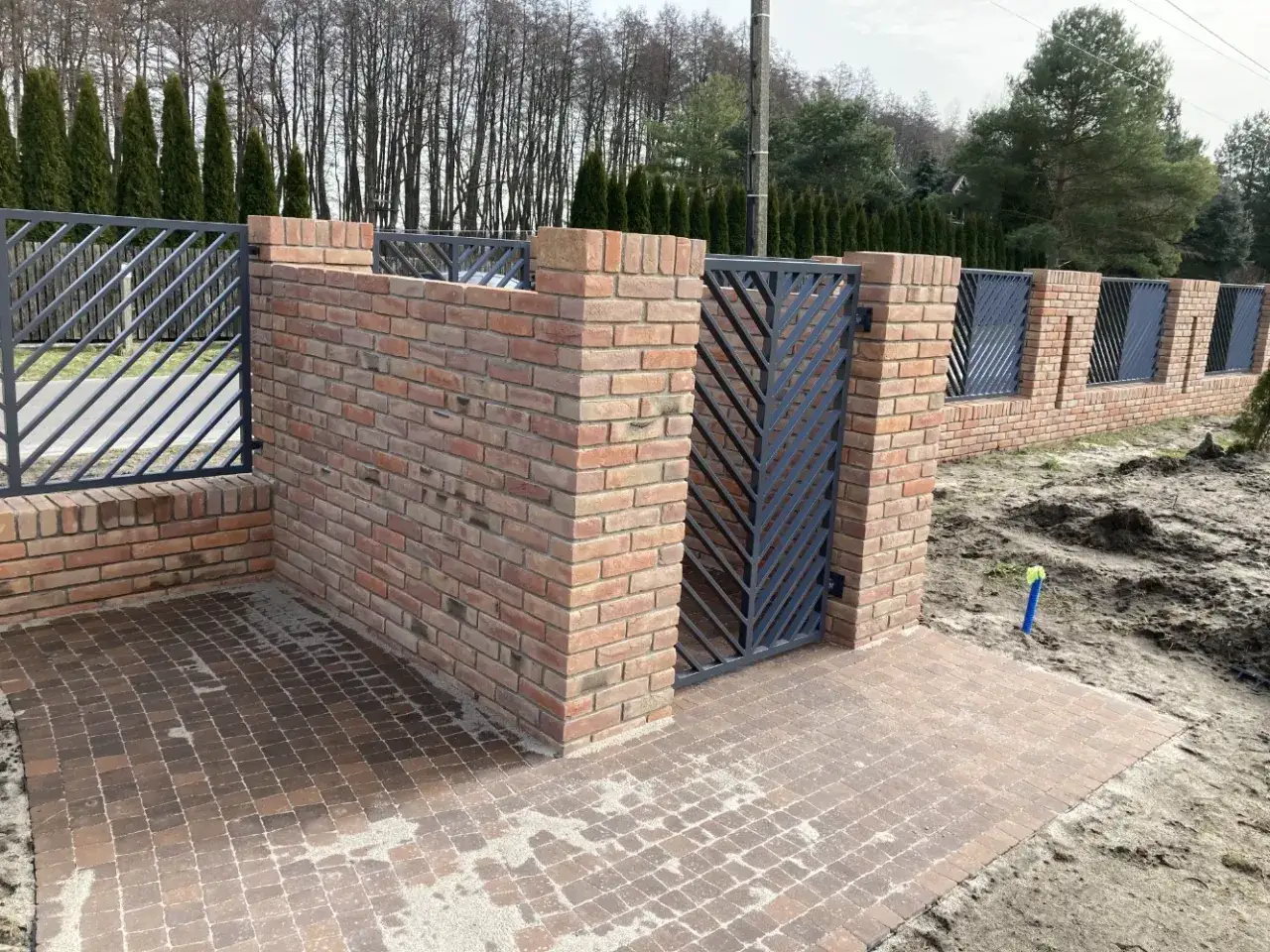 Nowe ogrodzenie z klinkieru z metalową bramą i furtką, otoczone brukowanym podjazdem i zielenią.