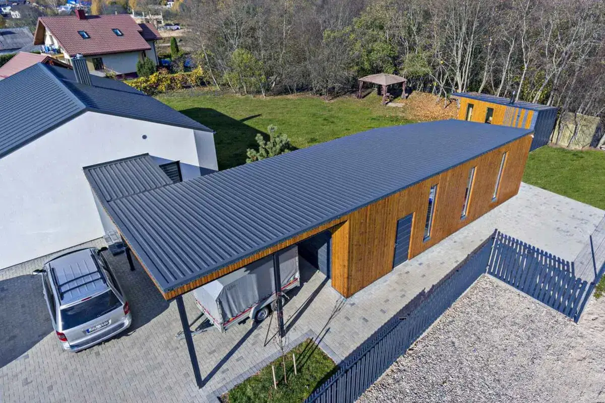 Nowoczesna konstrukcja dachu jednospadowego nad garażem i budynkiem gospodarczym. Elegancka, ciemnoszara blacha kryje przestronne, drewniane wnętrza.