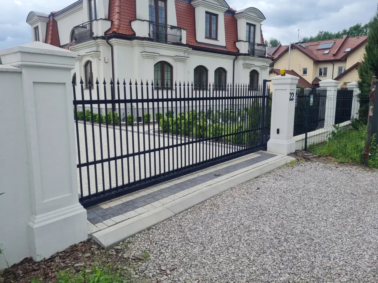 Eleganckie ogrodzenia w stylu dworkowym, z czarnej kutej stali, otaczają okazały biały dom z czerwoną dachówką.