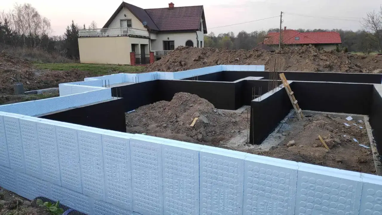 Fundamenty domu w budowie, izolowane styropianem. Widać czarne ściany fundamentowe i białe płyty izolacyjne.