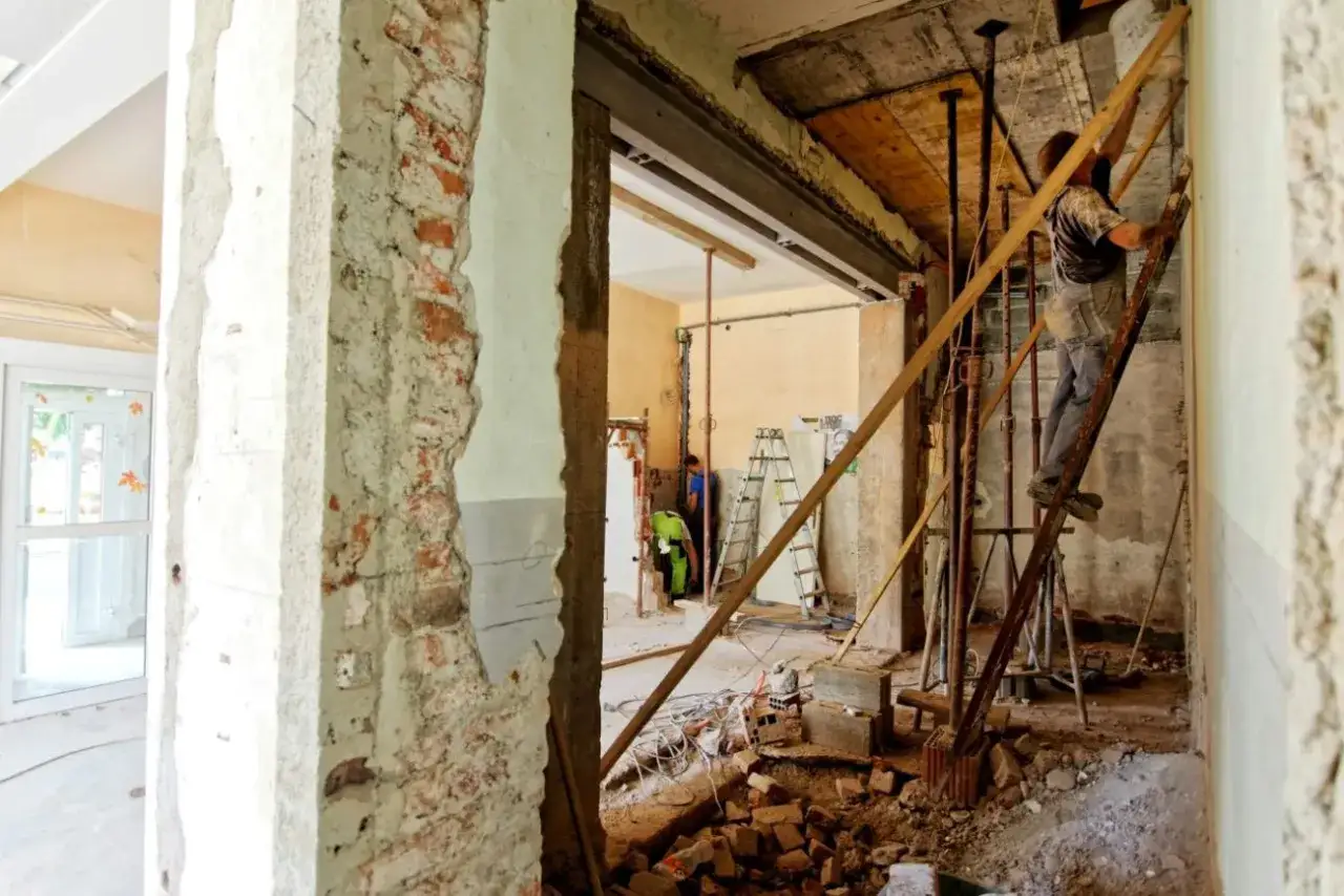 Remontowane wnętrze, gdzie widać odsłonięte cegły, betonowe słupy i rusztowania. Pracownicy zajmują się wzmacnianiem ścian nośnych i działowych.