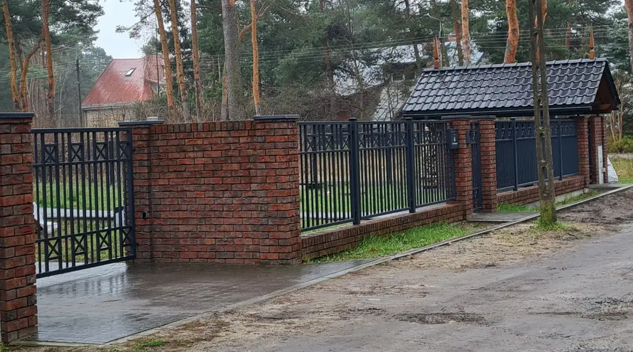 Nowoczesne ogrodzenie z cegły i czarnego metalu, z bramą wjazdową i zadaszeniem.