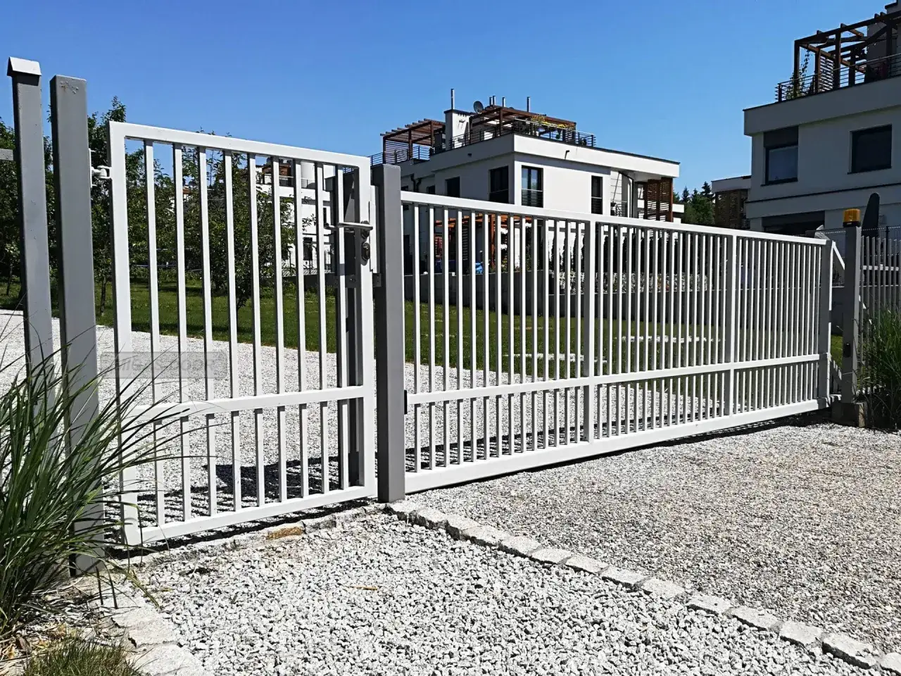 Brama przesuwna na pilota, biała, z pionowymi szczebelkami, otwiera wjazd na posesję z nowoczesnymi budynkami.
