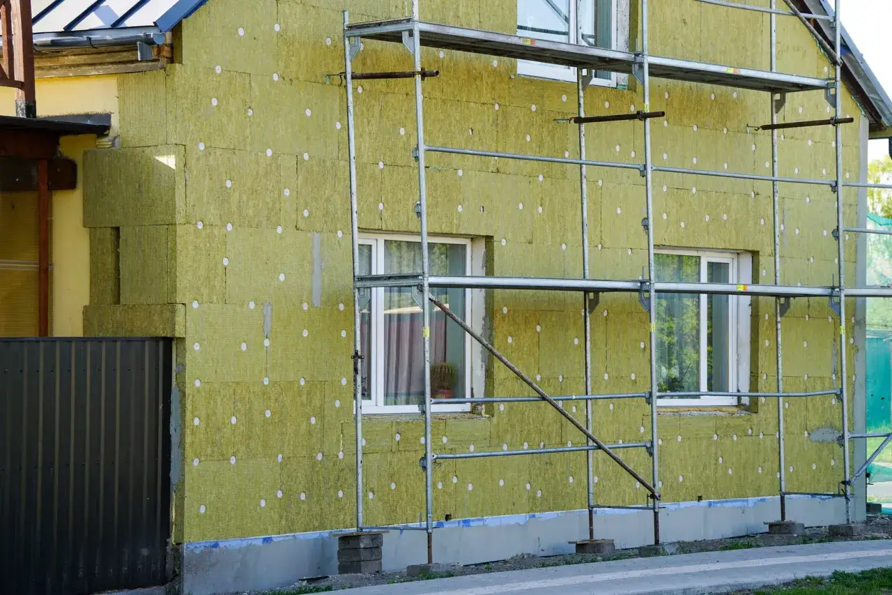 Dom w trakcie termomodernizacji, widoczne rusztowania i wełna mineralna na ścianach. Zewnętrzne glify okienne są częścią izolacji.