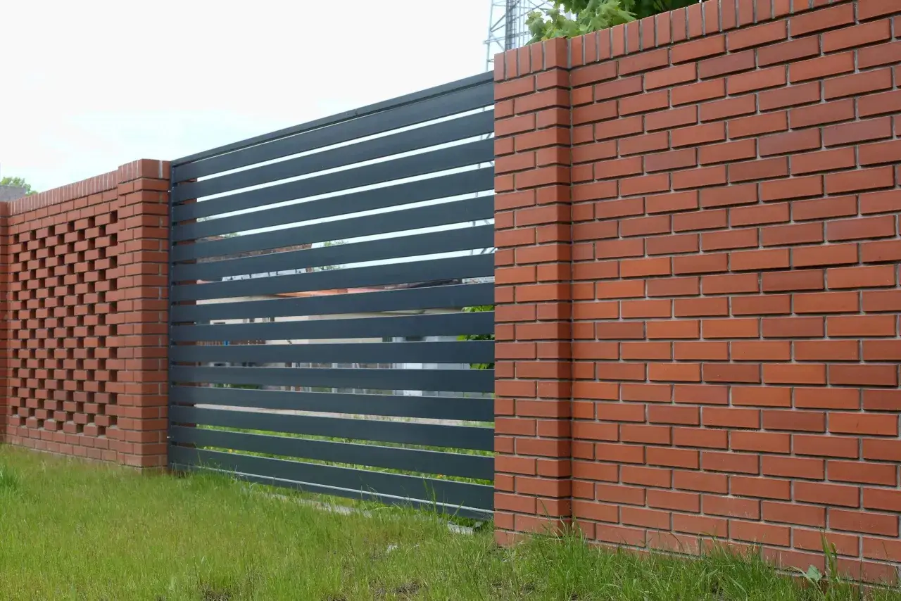Nowoczesne ogrodzenie z klinkieru i ciemnoszarej metalowej bramy. Ceglane słupki i ażurowa ściana dodają elegancji.