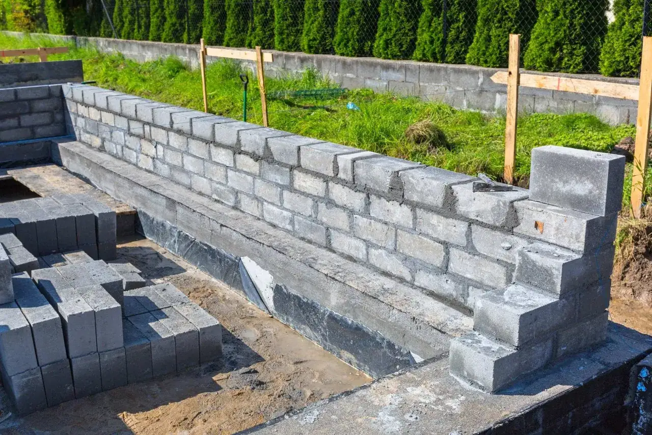 Budowa fundamentów: wznoszony mur z bloczka betonowego, stosy bloczków obok, w tle żywopłot i ogrodzenie.