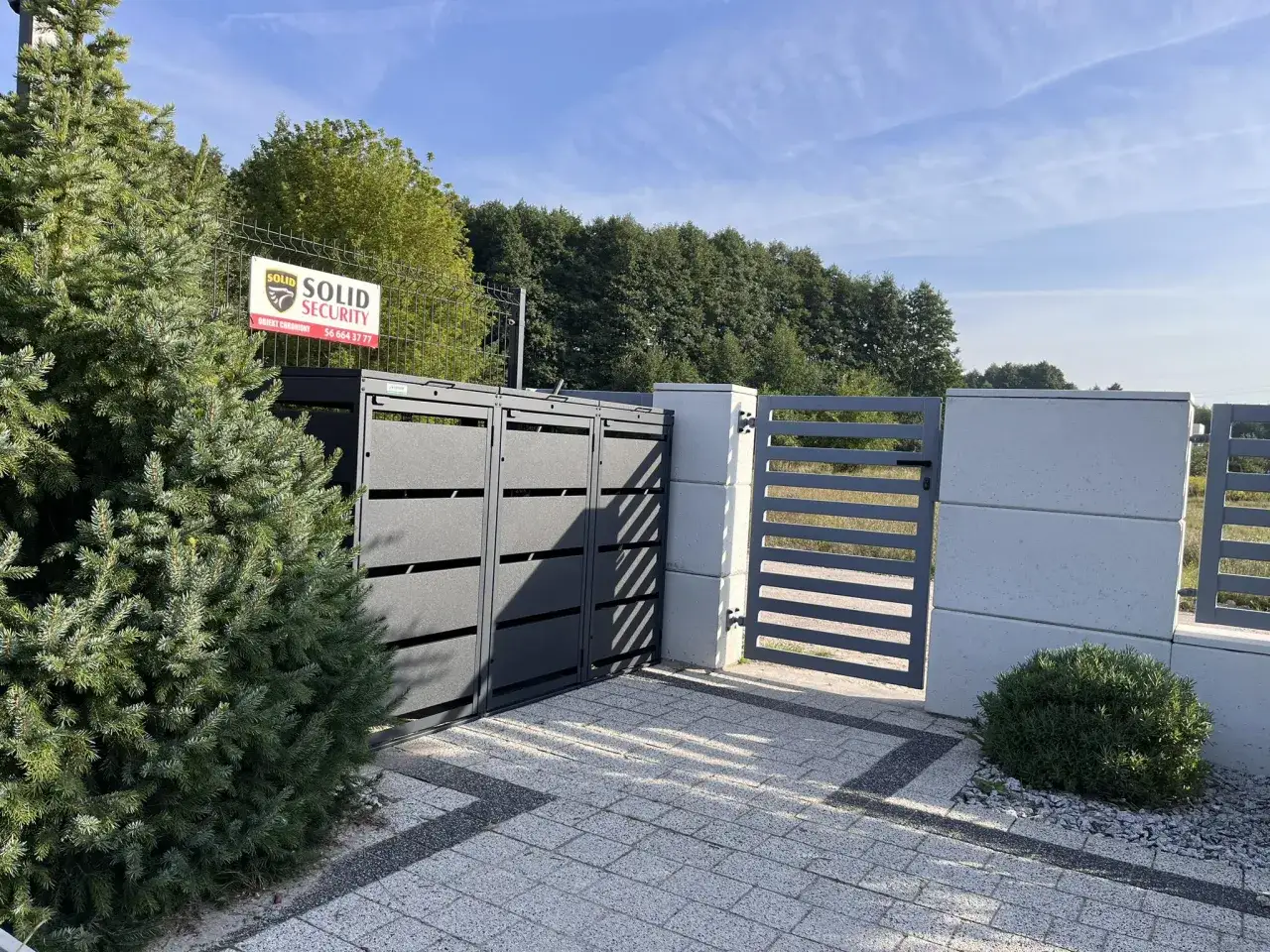 Nowoczesne śmietnik w ogrodzeniu, z szarymi panelami i betonowymi słupkami, otoczony zielenią.